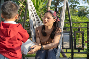 Obraz premium A young Asian mother and her young son spend the weekend together. Happily, outside the house, the background of bright and warm natural green trees. The concept of family ties.