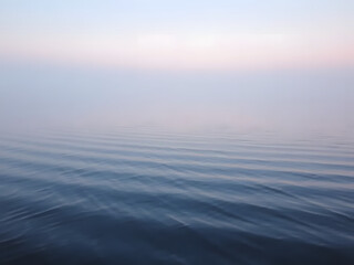 Fototapeta premium Gentle ripples on a calm lake at dawn, with fog and subtle pink and blue tones in the air