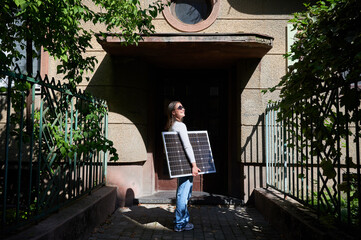 Woman holding solar panel for charging, stands in front of old building's entrance. Smiling girl wears sunglasses, white top, and blue jeans. Scene framed by green foliage and iron fencing.
