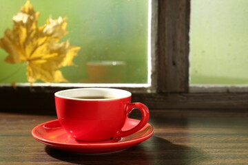 Red cup of aromatic coffee and saucer on wooden sill near window, closeup. Space for text