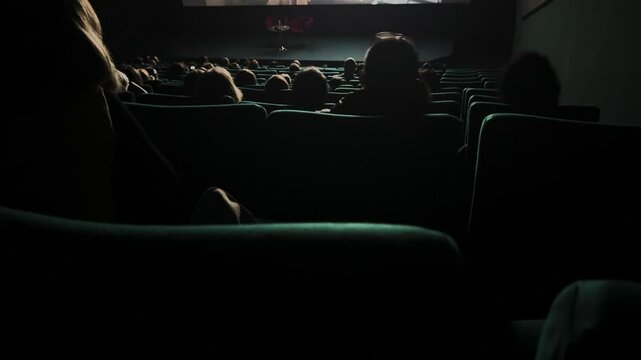 Audience dimly lit movie theater. Empty stage, anticipation. Plush teal seats. Film events premiere. People sit cinema watching at screen. Many spectators dark hall. Back rear view. Evening leisure.