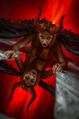 Beautiful angel woman in red lingerie with devilish horns and black wings looking in the mirror on a red background...
