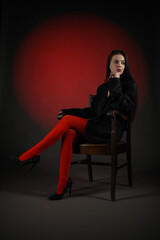 Beautiful girl in a black oversized blazer and red tights posing on the chair on a dark background.