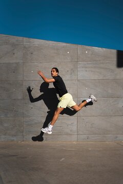 A man wearing yellow shorts and a black shirt leaps dynamically in the air, casting a shadow on a gray concrete wall under a bright blue sky