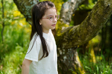 Little girl in glasses and T-shirt near tree in summer outdoors. Photo of happy active healthy child