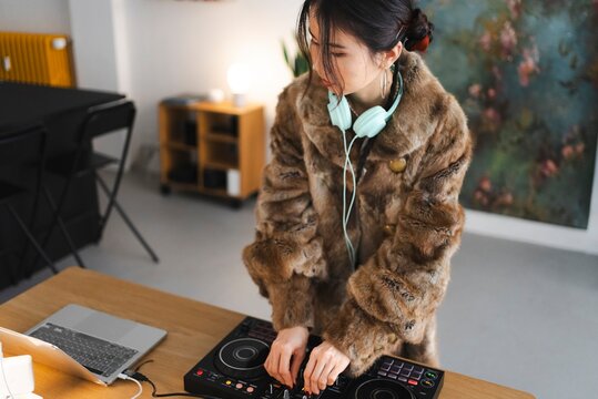 A female DJ wearing a fur coat adjusts the knobs on a DJ controller while wearing light blue headphones