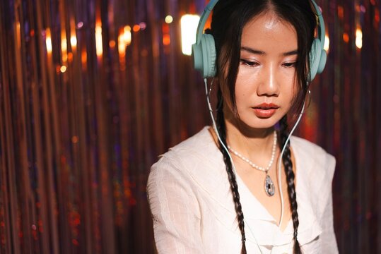 A focused female DJ in a white shirt and light blue headphones performs against a shimmering, colorful backdrop