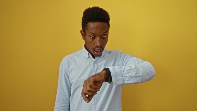Young african american man standing worriedly, wearing shirt and looking at watch over isolated yellow background, anxious about time and being late