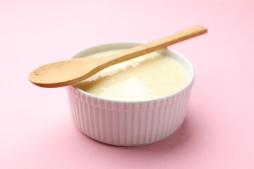 Tasty condensed milk in bowl and spoon on light pink background, closeup