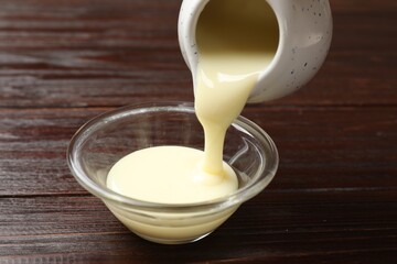 Pouring delicious condensed milk from jug into bowl on wooden table, closeup