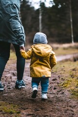 Obraz premium Small child wearing a yellow jacket and wool hat holding parent's hand while walking on a dirt path in the woods