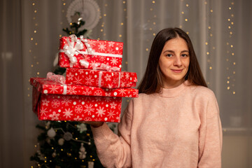 Smiling Woman Holding Christmas Gift in Light Pink Sweater by Decorated Tree, Cozy Holiday Scene with Twinkling Lights, Warm Winter Atmosphere, Festive Spirit, Joyful Christmas and New Year