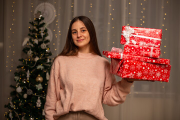 Smiling Woman Holding Christmas Gift in Light Pink Sweater by Decorated Tree, Cozy Holiday Scene with Twinkling Lights, Warm Winter Atmosphere, Festive Spirit, Joyful Christmas and New Year