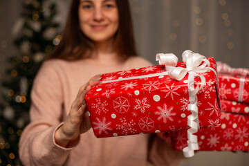 Smiling Woman Holding Christmas Gift in Light Pink Sweater by Decorated Tree, Cozy Holiday Scene with Twinkling Lights, Warm Winter Atmosphere, Festive Spirit, Joyful Christmas and New Year