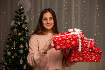 Smiling Woman Holding Christmas Gift in Light Pink Sweater by Decorated Tree, Cozy Holiday Scene with Twinkling Lights, Warm Winter Atmosphere, Festive Spirit, Joyful Christmas and New Year