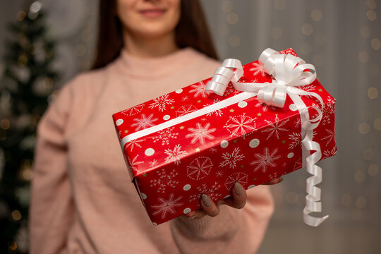 Smiling Woman Holding Christmas Gift in Light Pink Sweater by Decorated Tree, Cozy Holiday Scene with Twinkling Lights, Warm Winter Atmosphere, Festive Spirit, Joyful Christmas and New Year