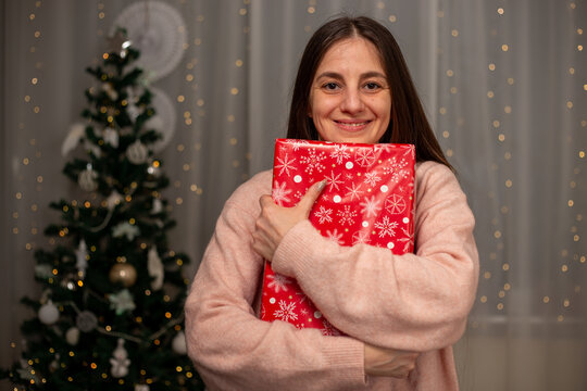 Smiling Woman Holding Christmas Gift in Light Pink Sweater by Decorated Tree, Cozy Holiday Scene with Twinkling Lights, Warm Winter Atmosphere, Festive Spirit, Joyful Christmas and New Year