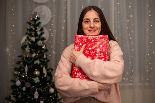 Smiling Woman Holding Christmas Gift in Light Pink Sweater by Decorated Tree, Cozy Holiday Scene with Twinkling Lights, Warm Winter Atmosphere, Festive Spirit, Joyful Christmas and New Year