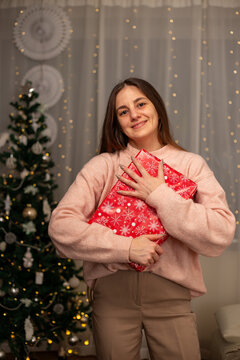Smiling Woman Holding Christmas Gift in Light Pink Sweater by Decorated Tree, Cozy Holiday Scene with Twinkling Lights, Warm Winter Atmosphere, Festive Spirit, Joyful Christmas and New Year