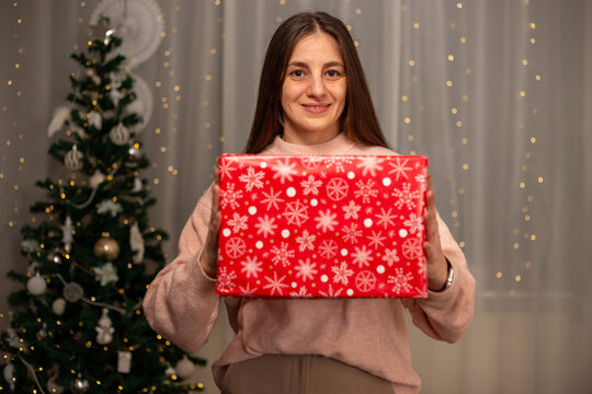 Smiling Woman Holding Christmas Gift in Light Pink Sweater by Decorated Tree, Cozy Holiday Scene with Twinkling Lights, Warm Winter Atmosphere, Festive Spirit, Joyful Christmas and New Year