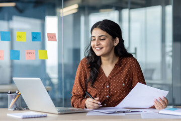 Smiling businesswoman seated at a desk, engaged in paperwork and using a laptop. Office setups and...