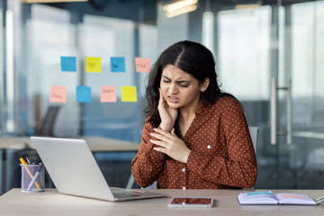A businesswoman at her desk in an office environment holds her face in discomfort, showcasing dental or health-related pain, possibly related to inflammation, while interacting with her work setup.