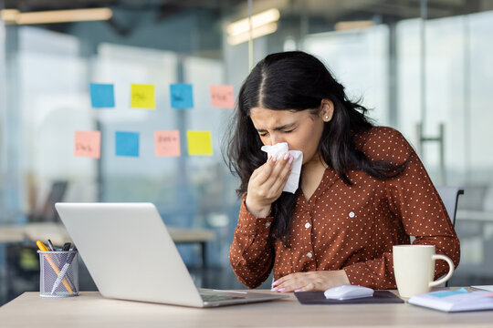 Businesswoman sneezing with a tissue at her office desk, surrounded by work materials and a laptop. Demonstrates topics such as illness, work-related stress, and the importance of health in workplace