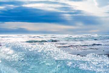 Wave crashing on black sand beach with iceberg breaking from glacier in sunny day