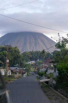 A peaceful village road leading to a towering mountain shrouded in clouds.