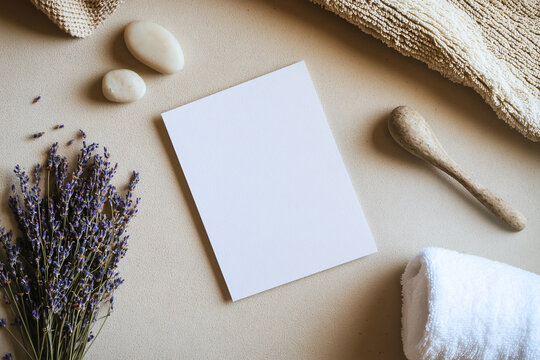 A blank invitation card rests on a matte beige surface surrounded by pebbles, dried lavender, and bathroom towels enhancing a serene atmosphere