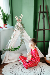 a funny boy in red pajamas sits with a soft toy in his hands on a knitted rug on the floor, against the backdrop of a children's wigwam