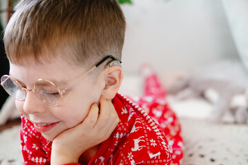 funny boy with hearing aids, glasses and red pajamas smiles widely while lying on a knitted rug on the floor