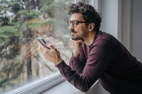 Young handsome middle eastern man scrolling on mobile phone in the morning standing near window.