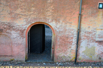 A weathered wall features a rounded archway, inviting visitors into a narrow pathway. The rustic...