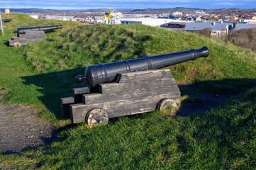 A historic cannon is positioned on grassy terrain, providing a stunning view of the city skyline under a clear blue sky. The scene captures a mix of nature and urban landscape