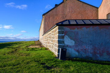 A historic building featuring a mix of stone and plaster walls stands amid lush green grass, illuminated by bright sunlight under a clear blue sky