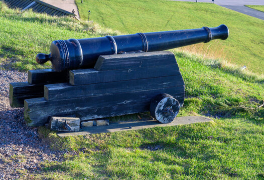 An old cannon is positioned on a grassy hill, offering a view of the shimmering coastline on a sunny afternoon. The structure is made of wood and metal, showcasing historical craftsmanship