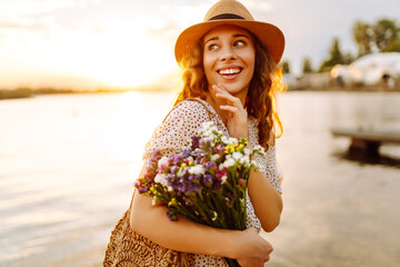 Happy woman in dress sitting on the wooden pier near the pond and clutched a bouquet of wildflowers tightly in her hand. Rustic style of the wedding.