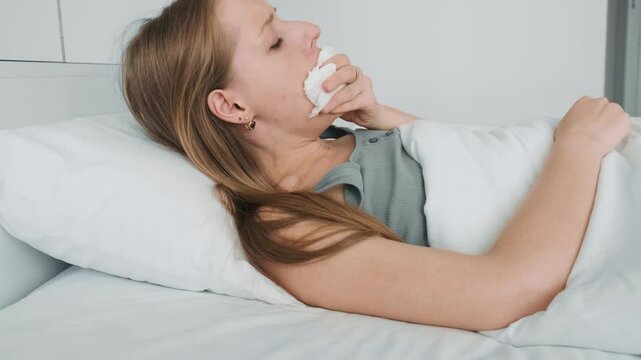 Sick woman with cold and flu, sneezing and coughing into tissue laying in bed. Infected girl with headache, blowing her nose and drinking medicine in a bedroom. Health care concept