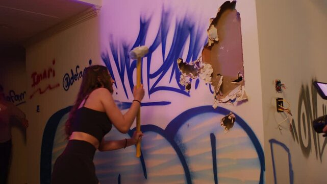 A girl swings and smashes a wall with a sledgehammer in the middle of a house party.