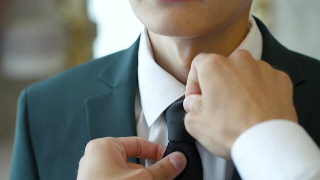 Male hands adjusting a black tie and collar on a neck in a white shirt and black neat jacket. Serving a customer in a man clothing store. Maintaining a business image. Concept of success and service - Powered by Adobe