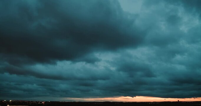 Timelapse of a wide angle view of the sky with fast moving dark blue clouds turning darker and getting an orange tint by city lights and of the horizon lit by sunset and lights of traffic on a highway