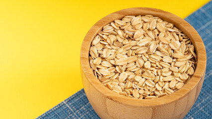Wooden bowl filled with rolled oats on a blue cloth, set against a bright yellow background. Perfect for healthy breakfast themes.
