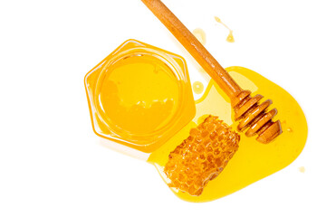 A hexagon glass jar with yellow sweet honey, honeycomb, and wooden honey dipper with leaked honey on a white background with copy space.