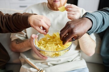 Front view closeup of diverse male hands grabbing potato chips from clear glass bowl during intense...