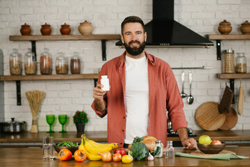 Smiling handsome caucasian man with beard in home kitchen holding bottle of nutritional supplements, looking at the camera. Painkiller, headache medication or vitamins concept