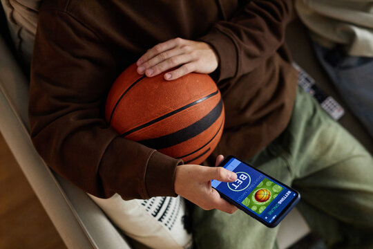 Top view of unrecognizable man holding basketball and smartphone with sports betting app while watching game at home, copy space