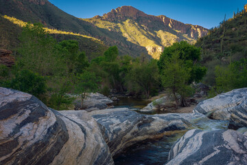 A serene wilderness scene showcasing a flowing stream surrounded by rugged desert terrain and green foliage, illuminated by golden sunrise rays over distant mountain peaks with a picturesque sky.