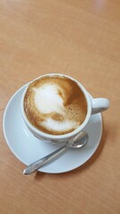 Xícara de café com leite, pires branco e colher, sobre mesa marrom.