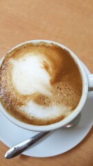 Close up de uma xícara de café com leite, pires branco e colher, sobre mesa marrom.